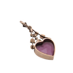Puede incluir: Colgante dorado con una gema morada facetada en forma de coraz&oacute;n. El colgante tiene un dise&ntilde;o floral decorativo encima del coraz&oacute;n y un peque&ntilde;o lazo en la parte superior para una cadena. El fondo es blanco.
