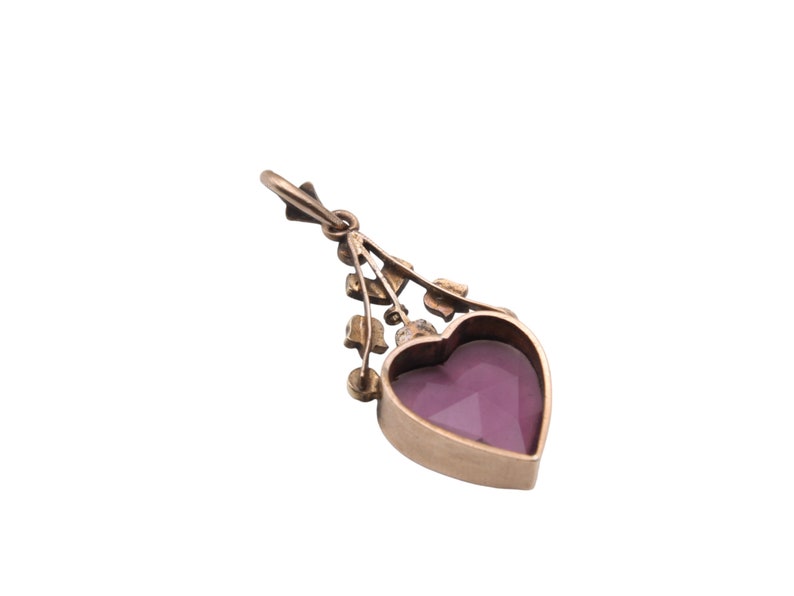 Puede incluir: Colgante dorado con una gema morada facetada en forma de coraz&oacute;n. El colgante tiene un dise&ntilde;o floral decorativo encima del coraz&oacute;n y un peque&ntilde;o lazo en la parte superior para una cadena. El fondo es blanco.