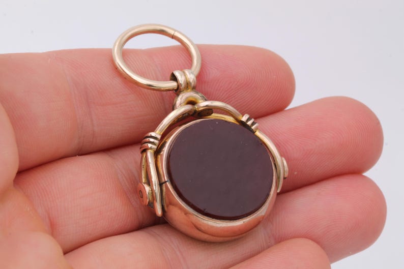 K&ouml;nnte beinhalten: Ein goldfarbener Anh&auml;nger mit einem kreisf&ouml;rmigen, dunkelroten Stein. Der Anh&auml;nger hat ein Drehdesign, wodurch sich der Stein drehen kann. Das Schmuckst&uuml;ck wird in einer Hand gehalten, um seine Gr&ouml;&szlig;e und Details zu zeigen.