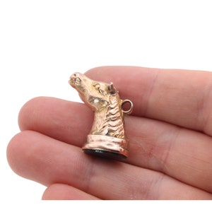 May include: A gold-colored horse head charm with a detailed mane and a small loop at the top. The charm is held in a hand against a white background. The horse head is facing upwards.