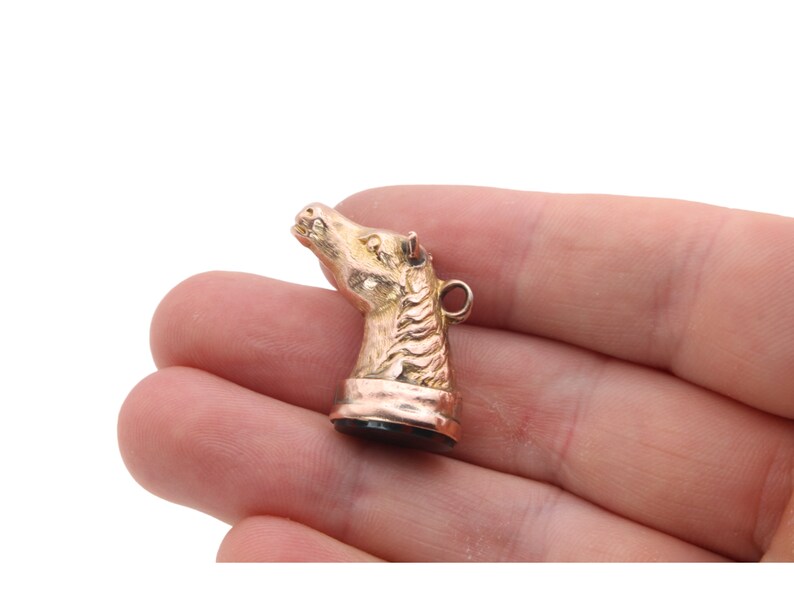 May include: A gold-colored horse head charm with a detailed mane and a small loop at the top. The charm is held in a hand against a white background. The horse head is facing upwards.