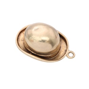 May include: A gold-colored charm in the shape of a bowler hat. The hat has a rounded crown and a curved brim. A small loop is attached for use as a pendant or charm. The charm is likely made of metal.