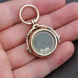 K&ouml;nnte beinhalten: Ein goldfarbener, runder Medaillon-Anh&auml;nger mit einer durchsichtigen Glasfl&auml;che, die einen kleinen, dunklen Innenraum freilegt. Das Medaillon hat ein Scharnierdesign und eine kleine Schlaufe zum Befestigen an einer Kette. Der Anh&auml;nger wird in einer Hand gehalten.