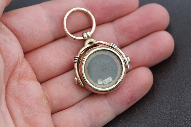 K&ouml;nnte beinhalten: Ein goldfarbener, runder Medaillon-Anh&auml;nger mit einer durchsichtigen Glasfl&auml;che, die einen kleinen, dunklen Innenraum freilegt. Das Medaillon hat ein Scharnierdesign und eine kleine Schlaufe zum Befestigen an einer Kette. Der Anh&auml;nger wird in einer Hand gehalten.