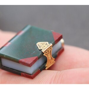 May include: Miniature book with a green cover and red corner accents. The book has a gold-colored clasp with an ornate design. The pages appear to be white. A small, detailed collectible item.