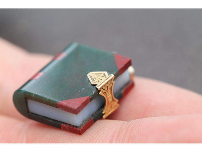 May include: Miniature book with a green cover and red corner accents. The book has a gold-colored clasp with an ornate design. The pages appear to be white. A small, detailed collectible item.