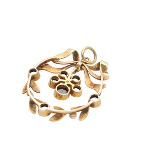 May include: A gold-toned pendant featuring a wreath design with a bow and a central flower-like element. The wreath is adorned with small, round gemstones and leaf details. The pendant is suspended from a small loop.