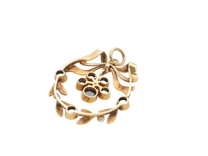 May include: A gold-toned pendant featuring a wreath design with a bow and a central flower-like element. The wreath is adorned with small, round gemstones and leaf details. The pendant is suspended from a small loop.