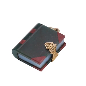May include: A miniature book charm with a dark green cover, red accents, and a gold-colored clasp. The charm has a detailed design and a small loop for attaching to a necklace or bracelet. The book's pages are a light color.
