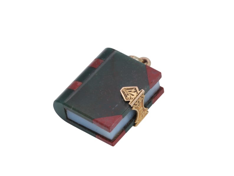 May include: A miniature book charm with a dark green cover, red accents, and a gold-colored clasp. The charm has a detailed design and a small loop for attaching to a necklace or bracelet. The book's pages are a light color.