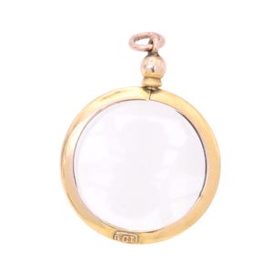 Op de afbeelding: Een goudkleurige hanger met een helder, cirkelvormig glazen oppervlak. De hanger heeft een kleine lus aan de bovenkant voor een ketting en een kleine rechthoekige markering aan de onderkant. Het ontwerp is eenvoudig en elegant.