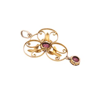 May include: A gold-toned pendant with a floral design, featuring a cluster of oval shapes and leaf accents. The pendant is adorned with several round, deep red gemstones. A single gemstone hangs from the bottom.