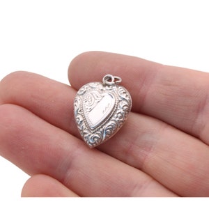 May include: A small, heart-shaped silver pendant with ornate scrollwork and a blank rectangular space for engraving. The pendant is held in a hand against a white background. The pendant is approximately 1 inch in length.