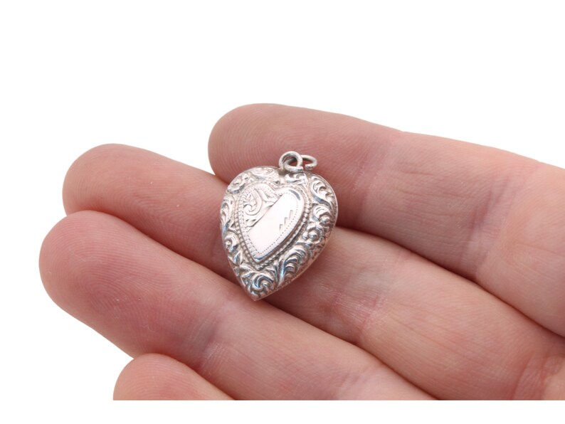 May include: A small, heart-shaped silver pendant with ornate scrollwork and a blank rectangular space for engraving. The pendant is held in a hand against a white background. The pendant is approximately 1 inch in length.