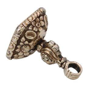 May include: Antique silver-toned seal with floral and scrollwork details. The seal features an oval face with embossed flowers and a decorative handle with a ring. A vintage piece, likely used for imprinting designs or initials.