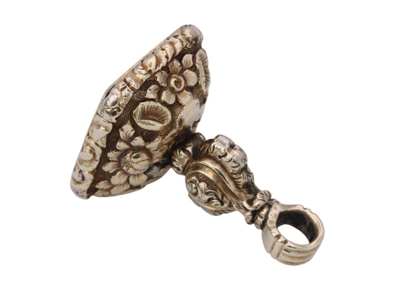 May include: Antique silver-toned seal with floral and scrollwork details. The seal features an oval face with embossed flowers and a decorative handle with a ring. A vintage piece, likely used for imprinting designs or initials.