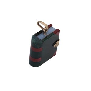 May include: A miniature book pendant with a gold-colored bail. The pendant features a dark green cover with red stripes and a gold-colored clasp. The top of the book is light blue and red.