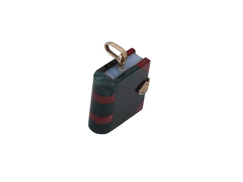 May include: A miniature book pendant with a gold-colored bail. The pendant features a dark green cover with red stripes and a gold-colored clasp. The top of the book is light blue and red.