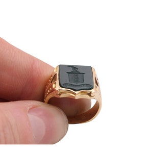May include: A gold signet ring featuring a rectangular black onyx inlay. The black stone is engraved with a crest design depicting a bird above a shield and a banner with text. The ring band has decorative cutouts along the sides of the shank.