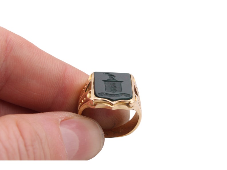 May include: A gold signet ring featuring a rectangular black onyx inlay. The black stone is engraved with a crest design depicting a bird above a shield and a banner with text. The ring band has decorative cutouts along the sides of the shank.