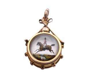 May include: A gold-toned pendant with a detailed miniature painting of a person riding a brown horse jumping over a barrier. The pendant has a round, glass-covered face and decorative accents.