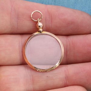 Op de afbeelding: Een goudkleurige, ronde hanger met een helder midden. De hanger heeft een kleine lus aan de bovenkant voor een ketting. De hanger wordt in de hand van een persoon gehouden.