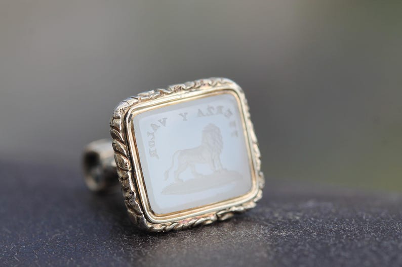 May include: A square ring with a gold-colored frame and a light-colored, possibly mother-of-pearl, center. The center features a lion engraving and text. The ring has a decorative, twisted band.