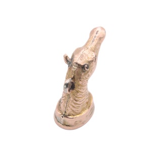 May include: A bronze-colored hippo figurine. The hippo's head is tilted upwards, with detailed features including ears, eyes, and textured skin. The figurine is mounted on a small, oval base. The overall design is detailed and artistic.