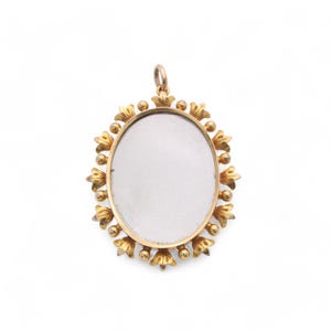 Op de afbeelding: Een goudkleurige ovale hanger met een helder midden. De hanger is omgeven door een decoratief bloemmotief. De hanger heeft een kleine lus aan de bovenkant voor een ketting.