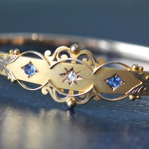 Könnte beinhalten: Goldfarbenes Armreif-Armband mit aufwendigen Details. Es hat drei blaue Edelsteine und einen zentralen sternförmigen Akzent mit einem klaren Edelstein. Das Armband hat ein filigranes Design und liegt auf dunklem blauem Hintergrund.