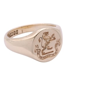 May include: A gold signet ring with a raised lion emblem. The ring has a smooth, polished surface and a rounded face. The lion is depicted in a heraldic pose, and the ring is set against a white background.