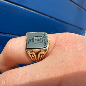 May include: A gold signet ring featuring a rectangular black stone inlay. The stone is engraved with a crest depicting a shield and a small figure. The ring band has a textured, beaded design along the sides, providing a detailed appearance.