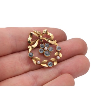 May include: A gold-toned pendant featuring a bow and a wreath design. The wreath is adorned with blue gemstones and small pearls. The pendant is held in a hand against a white background.