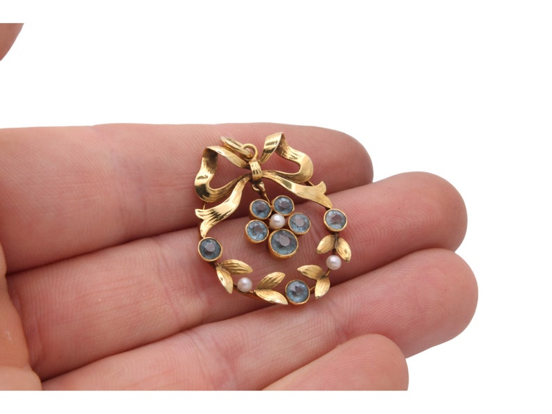 May include: A gold-toned pendant featuring a bow and a wreath design. The wreath is adorned with blue gemstones and small pearls. The pendant is held in a hand against a white background.