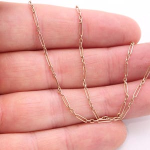 K&ouml;nnte beinhalten: Eine zarte, goldfarbene Kette mit einem sich wiederholenden rechteckigen Gliederdesign. Die Halskette wird in der Hand einer Person vor einem wei&szlig;en Hintergrund gehalten. Die Kettenglieder sind klein und gleichm&auml;&szlig;ig verteilt.