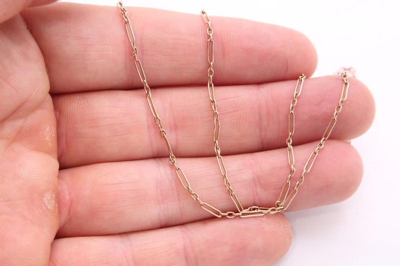 K&ouml;nnte beinhalten: Eine zarte, goldfarbene Kette mit einem sich wiederholenden rechteckigen Gliederdesign. Die Halskette wird in der Hand einer Person vor einem wei&szlig;en Hintergrund gehalten. Die Kettenglieder sind klein und gleichm&auml;&szlig;ig verteilt.