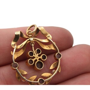 May include: A gold pendant with a wreath design. The pendant features a ribbon, leaves, and small, round, dark red stones. A flower-like charm hangs from the center. The pendant is held by a person's fingers.