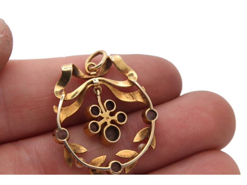 May include: A gold pendant with a wreath design. The pendant features a ribbon, leaves, and small, round, dark red stones. A flower-like charm hangs from the center. The pendant is held by a person's fingers.
