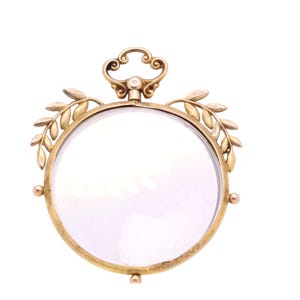 Op de afbeelding: Een goudkleurige loep met een heldere lens. Het ronde frame is versierd met bladeren en een decoratief handvat. De loep heeft een kleine, heldere edelsteen aan de bovenkant van het handvat.