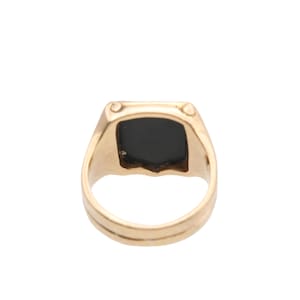 May include: A gold ring featuring a square-shaped black stone set into the centre of the band. The metal band has a smooth, polished finish and a flat top surface that holds the dark, opaque stone securely in place within the gold setting.