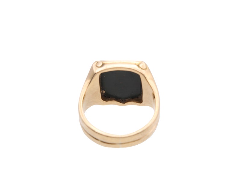 May include: A gold ring featuring a square-shaped black stone set into the centre of the band. The metal band has a smooth, polished finish and a flat top surface that holds the dark, opaque stone securely in place within the gold setting.