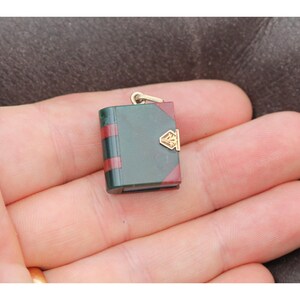 May include: A miniature book pendant with a gold-colored bail. The book has a dark green cover with red stripes and red edges. A gold-colored clasp adorns the front. The pendant is held in a person's hand.