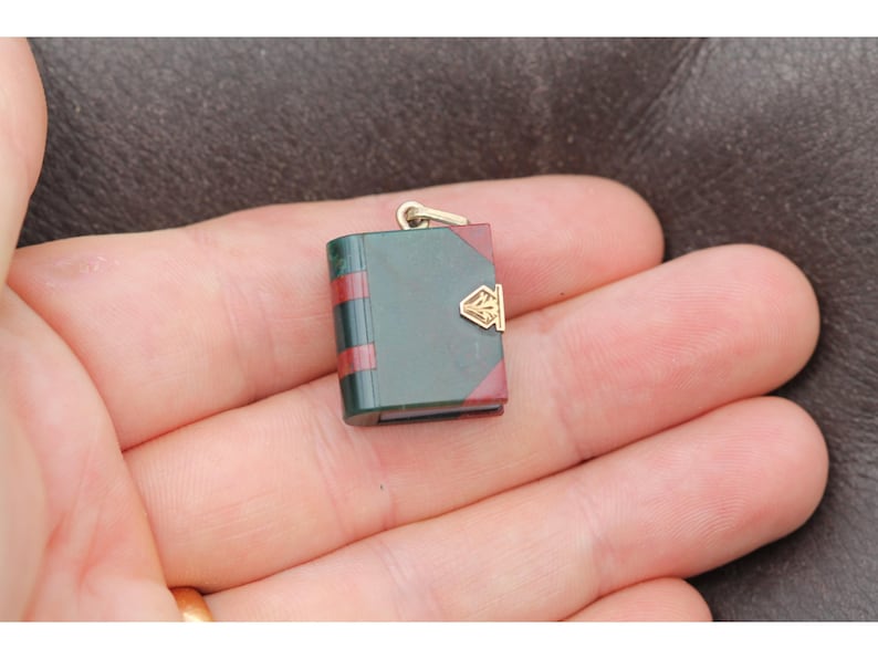 May include: A miniature book pendant with a gold-colored bail. The book has a dark green cover with red stripes and red edges. A gold-colored clasp adorns the front. The pendant is held in a person's hand.