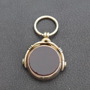 K&ouml;nnte beinhalten: Ein goldfarbener Anh&auml;nger mit einem kreisf&ouml;rmigen, dunkelroten Stein. Der Anh&auml;nger hat ein Drehdesign und einen kleinen Ring oben zum Anbringen an einer Kette. Der Stein ist in einen goldfarbenen Metallrahmen gefasst.
