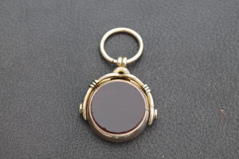 K&ouml;nnte beinhalten: Ein goldfarbener Anh&auml;nger mit einem kreisf&ouml;rmigen, dunkelroten Stein. Der Anh&auml;nger hat ein Drehdesign und einen kleinen Ring oben zum Anbringen an einer Kette. Der Stein ist in einen goldfarbenen Metallrahmen gefasst.
