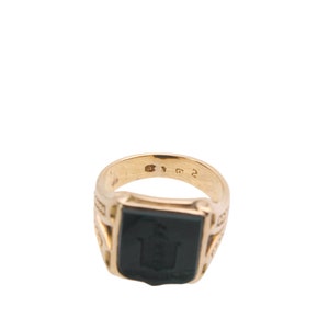 May include: A gold signet ring featuring a rectangular black onyx inlay with an engraved crest design. The interior of the band displays clear hallmarks and numerical stamps, showcasing a polished metallic finish and a structured, solid design.
