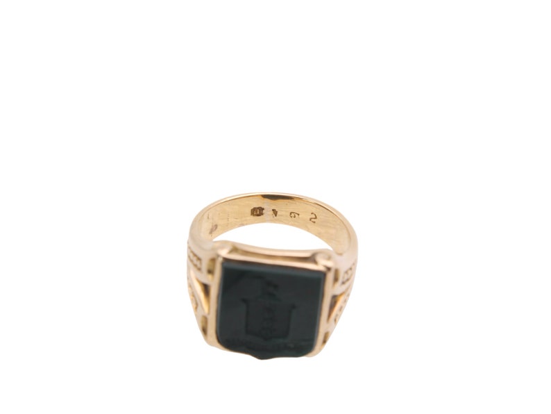 May include: A gold signet ring featuring a rectangular black onyx inlay with an engraved crest design. The interior of the band displays clear hallmarks and numerical stamps, showcasing a polished metallic finish and a structured, solid design.