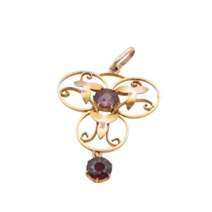 May include: A gold-toned pendant with a floral design, featuring three circular elements and a dangling gemstone. The pendant has three purple gemstones and a small loop at the top for a chain. The pendant is set against a white background.