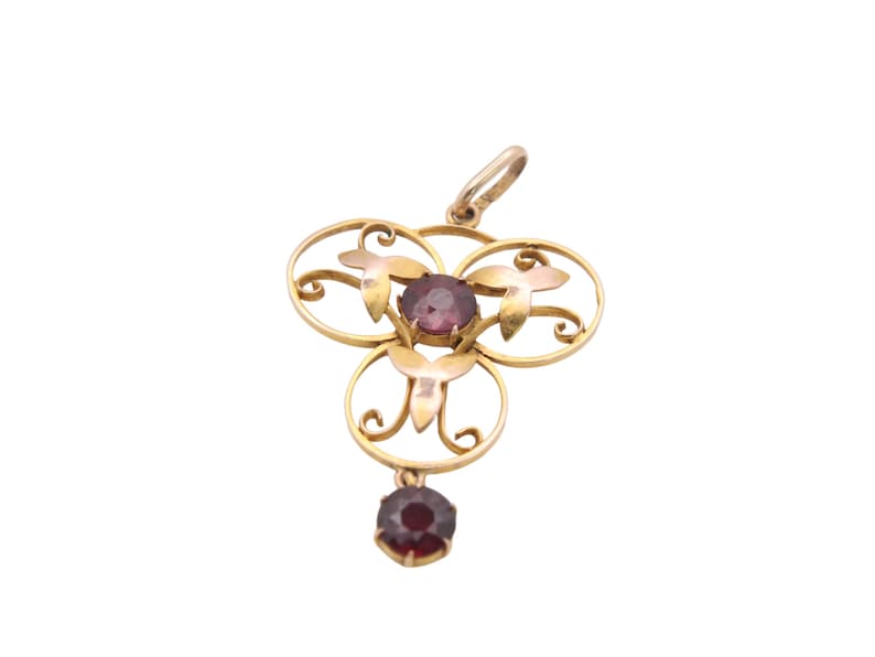 May include: A gold-toned pendant with a floral design, featuring three circular elements and a dangling gemstone. The pendant has three purple gemstones and a small loop at the top for a chain. The pendant is set against a white background.