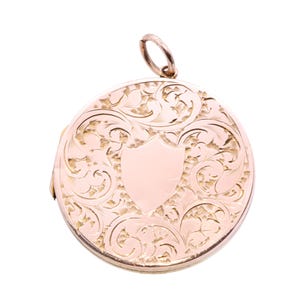 May include: A round, gold-colored locket with intricate floral and scroll engraving. A blank shield shape is centered on the front. The locket has a small loop at the top for a chain.